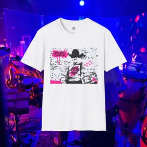 May include: White t-shirt featuring a graphic design with a person wearing a cowboy hat, musical notes, and the text "HELLO DENNY" in pink. The design is in black and white with pink accents.