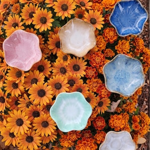 Könnte beinhalten: Sechs kleine, blütenförmige Keramikschalen in verschiedenen Farben: Rosa, Beige, Blau, Hellblau, Grün und Weiß. Die Schalen sind auf einem Bett aus leuchtend orangefarbenen Blumen mit dunkelbraunen Zentren angeordnet.