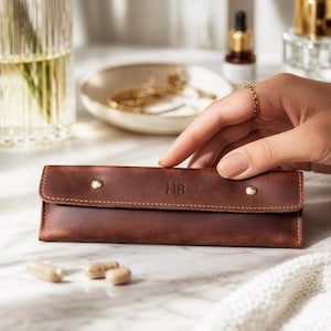Könnte beinhalten: Ein braunes Lederetui mit den Initialen "HB" auf der Vorderseite. Das Etui wird mit zwei goldfarbenen Knöpfen verschlossen. Mehrere Kapseln liegen vor dem Etui. Das Etui liegt auf einer Marmoroberfläche.