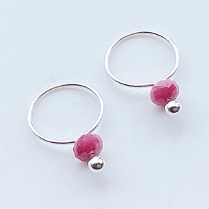 May include: A pair of silver hoop earrings. Each earring features a faceted, deep pink gemstone and a small silver bead. The earrings are set against a white background, highlighting their simple elegance and vibrant color.