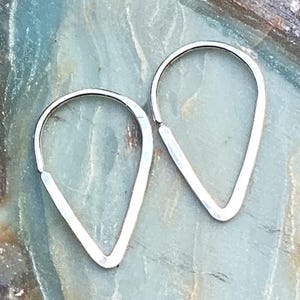 May include: A pair of silver teardrop-shaped earrings. The earrings are made of a thin, polished metal and have a simple, minimalist design. The earrings are displayed on a blue and green marbled surface.