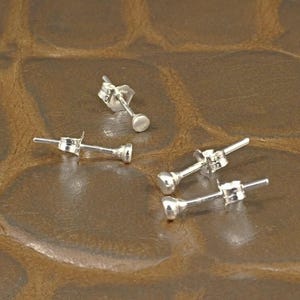 Peut inclure: Quatre petites boucles d'oreilles à clous argentées avec une tige et un devant rond et plat. Les boucles d'oreilles sont éparpillées sur une surface marron texturée. Les boucles d'oreilles sont probablement en argent sterling.