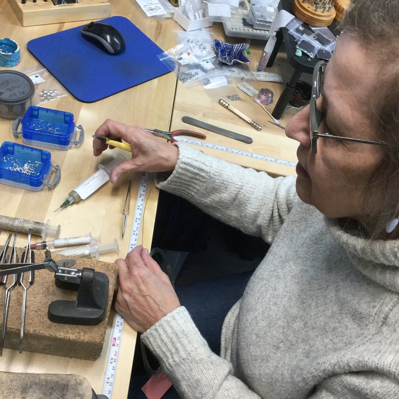 Puede incluir: Un taller de fabricaci&oacute;n de joyas con herramientas, peque&ntilde;os recipientes, jeringas y una cinta m&eacute;trica. Una persona con un su&eacute;ter gris claro y gafas trabaja en el banco.