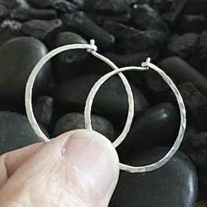 May include: A pair of silver hoop earrings with a hammered texture. The earrings are round and have a simple, classic design. The hoops are held by a person's hand against a dark background.