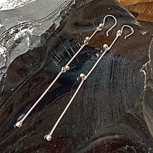 May include: A pair of long, silver earrings with small, round beads. The earrings are suspended from a simple hook.