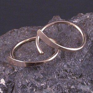 Puede incluir: Dos pendientes de aro dorados con un dise&ntilde;o &uacute;nico e entrelazado. Los pendientes se muestran sobre una superficie oscura y texturizada, mostrando su estilo elegante y minimalista. Los aros tienen un acabado liso y pulido.