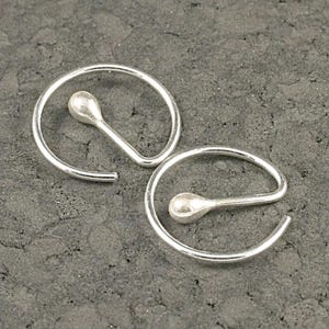 May include: A pair of silver hoop earrings. Each earring has a teardrop-shaped charm attached to a curved, open hoop. The earrings are shown against a dark grey textured surface.