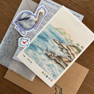 Pu&ograve; includere: Una collezione di articoli di cancelleria tra cui una cartolina acquerellata di La Jolla, un adesivo con un pellicano e delle buste. La cartolina presenta una scena costiera con il testo "la jolla". Una busta ha un'illustrazione di foca e la scritta "handle with care".