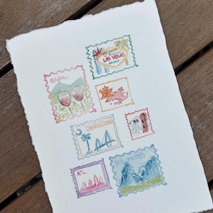 Peut inclure: Peinture &agrave; l'aquarelle de timbres-poste repr&eacute;sentant des destinations de voyage. Les timbres incluent Napa, Las Vegas, Charleston et les Dolomites. L'&oelig;uvre est sur papier blanc textur&eacute; avec un bord d&eacute;coup&eacute;, sur fond de bois.