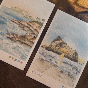 May include: Two watercolor paintings of coastal scenes. One depicts a rocky coastline with crashing waves and the other shows a rock formation with a natural arch. The text "big sur" is printed on one of the paintings.