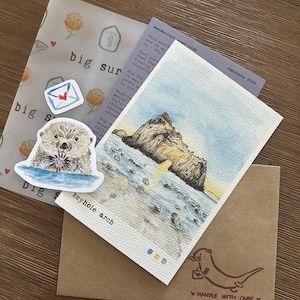 May include: A collection of Big Sur themed stationery. Includes a watercolor print of Keyhole Arch, a sticker of an otter, and a brown envelope with an otter illustration and the text "Handle with Care". Also includes a translucent sheet with the text "Big Sur".