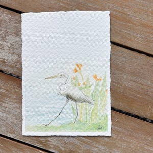 May include: Watercolor painting of a white heron wading in shallow water, with green reeds and orange flowers in the background. The artwork is on textured paper with a deckled edge. Signed in the lower right corner.