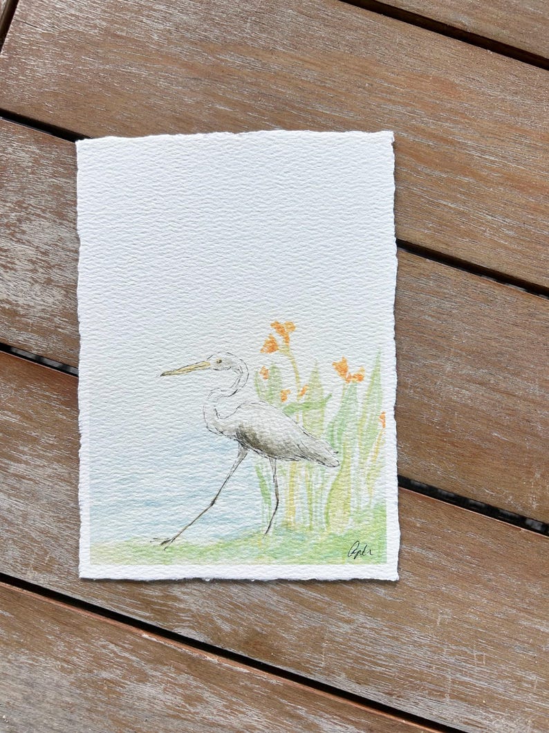 May include: Watercolor painting of a white heron wading in shallow water, with green reeds and orange flowers in the background. The artwork is on textured paper with a deckled edge. Signed in the lower right corner.