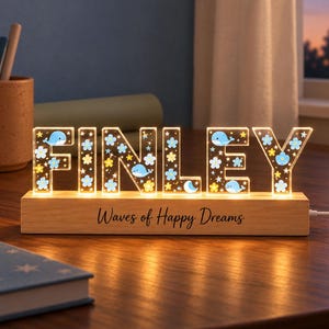 May include: A light-up sign spelling FINLEY in clear acrylic letters decorated with blue whales, yellow stars, and blue flowers. The letters sit on a rectangular wooden base with the text Waves of Happy Dreams printed in black.