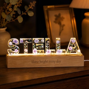 Op de afbeelding: Een verlicht display van heldere hars die de naam STELLA vormt, met ingebedde blauwe en paarse gedroogde bloemen. De letters staan op een rechthoekige houten voet met de gegraveerde tekst Shine bright every day, verlicht door een interne lichtbron.