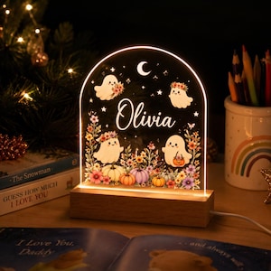 May include: An arched acrylic night light with a wooden base, featuring the name Olivia in white script. The design includes four cute ghosts, colorful flowers, pumpkins, stars, and a crescent moon, all illuminated with a warm glow.