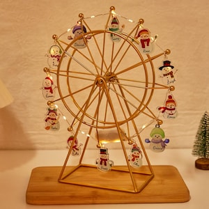 Peut inclure: Une décoration de Noël en forme de grande roue dorée avec des ornements de bonhommes de neige, chacun portant un nom. La roue est illuminée par de petites lumières et repose sur une base en bois. Un petit sapin de Noël est en arrière-plan.