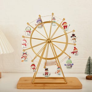 Peut inclure: Décoration de Noël en forme de grande roue dorée avec des ornements de bonhommes de neige. Chaque ornement porte un nom, comme "David" et "Tommy". La roue repose sur une base en bois et est éclairée par de petites lumières.
