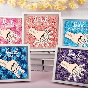 May include: A collection of framed floral art pieces with the words "Dad, We Love You" and a hand graphic. Each frame features a different color of roses: pink, peach, blue, dark blue, and purple. The hand graphic holds tags with names.