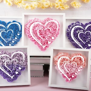 Può includere: Cinque composizioni floreali a forma di cuore incorniciate in tonalità di blu, rosa, viola e pesca. Ogni cuore è riempito di piccole rose e presenta un contorno a forma di cuore bianco con testi come "Mamma" e "Nonna".