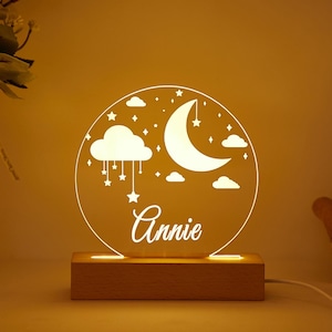 Puede incluir: Una luz nocturna de acrílico blanco con un diseño de luna, estrellas y nubes. La luz tiene una base de madera y el nombre "Annie" está impreso en la parte delantera.