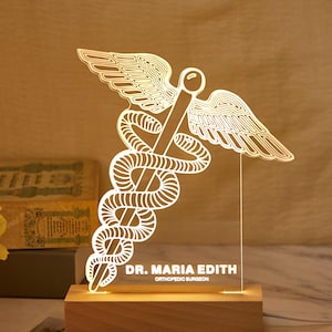 May include: A lit acrylic lamp displaying the Caduceus symbol, a staff with entwined serpents and wings. It sits on a wooden base. The lamp has the text "DR. MARIA EDITH ORTHOPEDIC SURGEON".