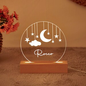 Puede incluir: Una luz nocturna circular de acrílico transparente con un diseño grabado en blanco de una luna creciente, estrellas y una nube colgando de líneas. El nombre Rocco está escrito en letra cursiva blanca bajo la luna sobre una base de madera.