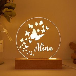 May include: A personalized night light featuring a circular design with a butterfly motif and the name "Alina" in white script. The light sits on a wooden base and emits a warm glow.