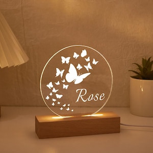 May include: A round, clear acrylic night light with a butterfly design and the name "Rose" etched in white. The light sits on a rectangular wooden base. A small potted plant and a lamp are in the background.