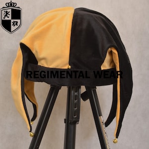 May include: A two-toned jester hat in black and gold velvet, with long, pointed tips adorned with small gold bells. The hat is displayed on a black stand, with the text "REGIMENTAL WEAR" visible.