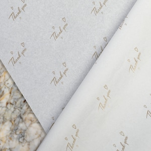 May include: White tissue paper with a repeating pattern of gold hearts and the words "Thank you".