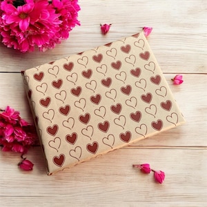 Puede incluir: Una bolsa de regalo de papel marrón con un patrón repetido de corazones rojos. La bolsa está sobre una superficie de madera clara con flores rosas esparcidas alrededor.
