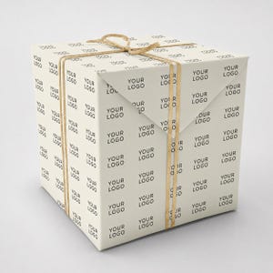 Può includere: Una scatola regalo quadrata avvolta in carta color crema con un motivo testuale nero ripetuto che recita YOUR LOGO. La scatola è fissata con uno spago sottile color marrone chiaro legato in un semplice fiocco sulla parte superiore, ben definita.