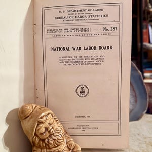 Puede incluir: Un libro antiguo abierto titulado "National War Labor Board" con texto del Departamento de Trabajo de EE. UU. Una figura de gnomo de cerámica descansa frente al libro. El libro muestra texto y un sello gubernamental.