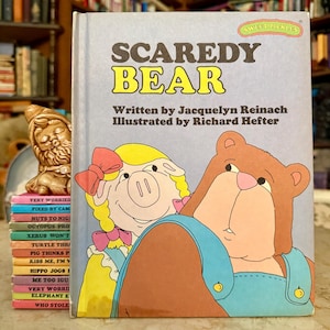 Puede incluir: Un libro infantil titulado "Scaredy Bear" con ilustraciones de un oso y un cerdo. La portada es azul claro con texto amarillo y marrón. El libro forma parte de la serie "Sweet Pickles". Se ve una pila de otros libros.