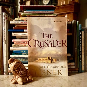 Puede incluir: Una novela titulada "The Crusader" con una portada en tonos sepia que presenta una ilustración de un castillo. El libro está apoyado entre pilas de otros libros y una pequeña figura de gnomo. El texto "Advance Reading Copy Not For Sale" es visible.