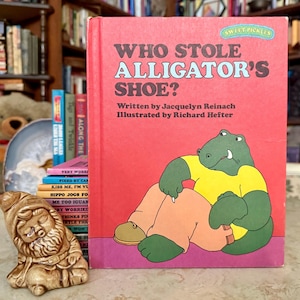 May include: A children's book titled "Who Stole Alligator's Shoe?" with a red cover and a cartoon alligator illustration. The book is part of the "Sweet Pickles" series. A small ceramic gnome figurine and a stack of other books are in the background.