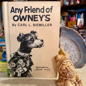 May include: A vintage book titled "Any Friend of Owney's" with a dog illustration on the cover. The book is accompanied by a small ceramic gnome figurine. The book's cover also includes the author's name and the illustrator's name.