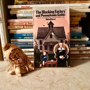 Puede incluir: Un libro de bolsillo vintage titulado "The Blacking Factory and Pennsylvania Gothic" de Wilfrid Sheed, con una ilustración de una casa. Una pequeña figura de gnomo marrón claro está frente al libro, con una pila de libros en el fondo.