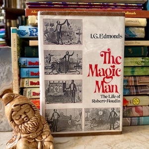 May include: A vintage book titled "The Magic Man" by I.G. Edmonds, with illustrations of a magician. The cover is white with red lettering. A ceramic gnome figurine sits in front of the book, with a stack of other books in the background.