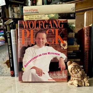 May include: A collection of books, including "Fear of Flying" and "The Story of the Stone", is stacked behind a cookbook titled "Wolfgang Puck Adventures in the Kitchen". A small gnome figurine is on the right.