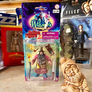 May include: An action figure of a wizard in a pink robe with a long beard, staff, and sword, packaged in a clear plastic case. The packaging reads "The Wizard's Magic" and "Action Figure". A small ceramic gnome is in the foreground.