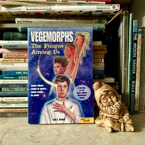 Può includere: Una copia vintage di "Vegemorphs: The Fungus Among Us" di Leif E. Green, con una copertina blu e illustrazioni di giovani. Una piccola statuina di gnomo in ceramica è a destra del libro, con una pila di libri sullo sfondo.