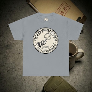 May include: A light blue t-shirt featuring a circular graphic with the text "CERTIFIED MONKEY BUSINESS - AGENCY OPERATIVE" and "EST. 2025". The graphic includes a hand holding a magnifying glass over a banana. The words "TOP SECRET" are also included.