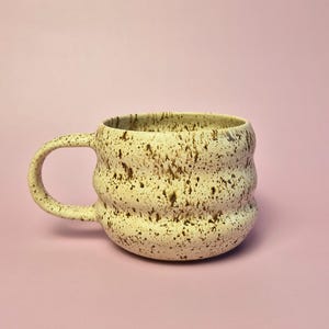 Könnte beinhalten: Cremefarbene Keramiktasse mit einem gesprenkelten braunen Muster. Die Tasse hat einen abgerundeten Körper mit gerippter Textur und einem großen, gebogenen Henkel. Die Tasse steht vor einem weichen rosa Hintergrund.