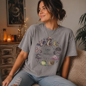Puede incluir: Una camiseta gris de manga corta con un estampado gráfico circular de varios animales, incluyendo un murciélago, una serpiente, una araña, una rana y un caracol, rodeando el texto "LOVE THE UNLOVED" con pequeños símbolos rúnicos.