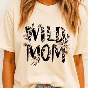 May include: Cream-colored t-shirt with the words "WILD MOM" in a black, leopard print font. Floral accents in shades of brown and white surround the text. The shirt is a casual style.