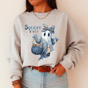 Op de afbeelding: Een lichtgrijs sweatshirt met een grillig spookontwerp en de tekst "Spooky Vibes". De geest is versierd met een bloemenstrik en draagt een pompoenmand. Het ontwerp gebruikt blauwtinten en bloempatronen, perfect voor Halloween.