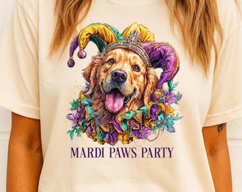 May include: Cream-colored t-shirt featuring a Golden Retriever wearing a jester hat and a floral collar. The dog has its tongue out and is smiling. The text "MARDI PAWS PARTY" is printed below the dog in purple.