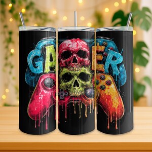 Puede incluir: Vaso negro con un diseño colorido con la palabra "GAMER" y un gráfico de calavera. El diseño incluye mandos de juego en rojo, azul y amarillo. El vaso tiene una pajita y tapa plateadas.